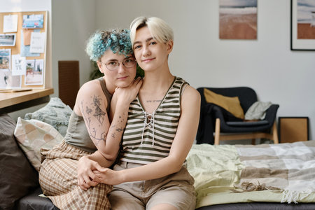 Portrait of lesbian girls looking at camera while sitting on bed in bedroomの写真素材