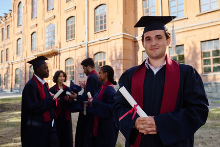 Portrait of young man with diploma looking at camera while standing in robe outdoorsの写真素材