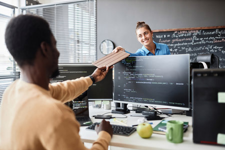 Woman giving tablet pc to her colleague while he sitting at his workplace and working with computer codesの写真素材