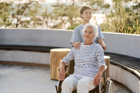Portrait of senior woman sitting in wheelchair and looking at camera while walking with nurse outdoorsの写真素材