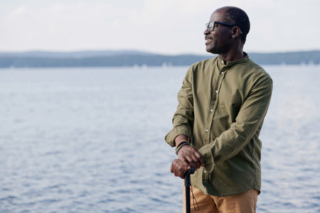 African American senior man with stick standing outdoors and looking at beautiful view of lake