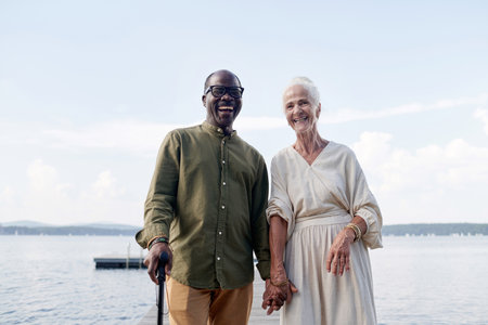 Portrait of multiethnic happy senior couple holding hands and smiling at camera while standing...