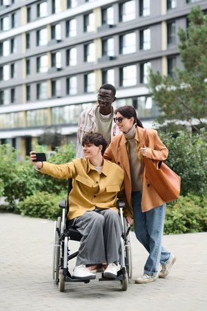 Vertical image of man with disability making selfie with his friends on smartphone during their walk...