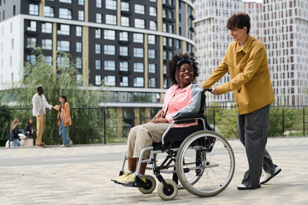 Multiethnic couple enjoying time outdoors, young man riding his friend in wheelchair along the streetの写真素材