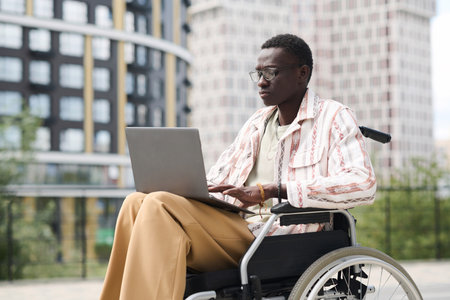 African American man with disability sitting on wheelchair and typing on laptop outdoorsの写真素材