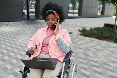 African American woman with disability having conversation on the phone while working online on laptopの写真素材