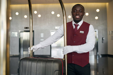 Portrait of African American doorman smiling at camera while standing with trolley for luggageの写真素材