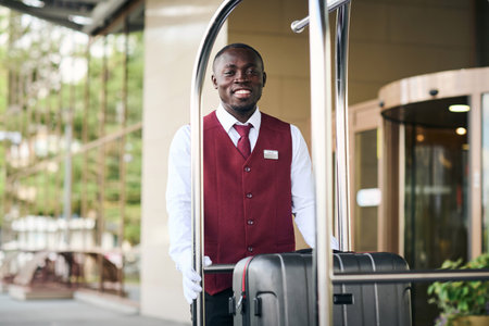 Portrait of African American doorman standing outdoors near the trolley with baggage and smiling at cameraの写真素材