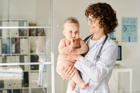Smiling doctor in white coat holding baby on arms while standing at officeの写真素材