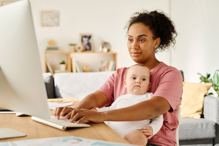 Mother working online on computer while sitting with baby at table in front of monitorの写真素材