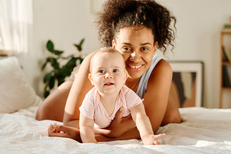 Portrait of happy mother smiling at camera while playing with her baby in bedroomの写真素材