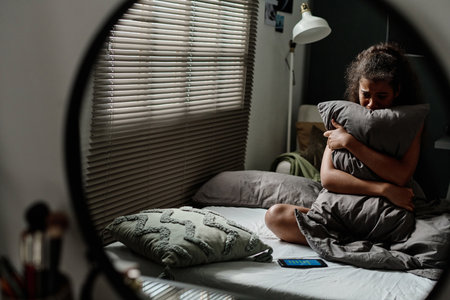 Reflection in mirror of crying female teenager with pillow sitting on double bed by window and...