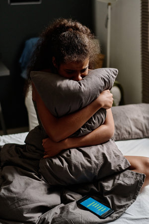 Miserable teenage girl crying in pillow while sitting on double bed with smartphone at night and suffering from cyberbullying messagesの写真素材