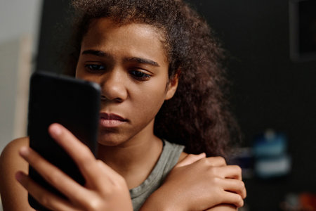 Stressed youthful girl with smartphone reading cyberbullying messages while standing in front of camera late at night in home environmentの写真素材