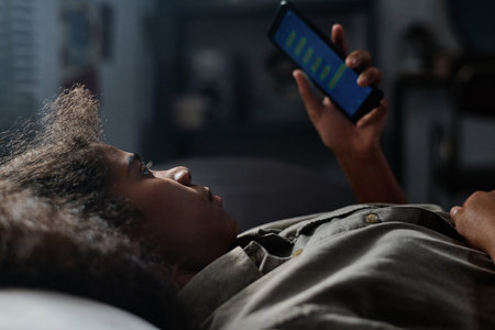 Side view of depressed female teenager looking through cyberbullying messages in her mobile phone while lying in bed late at nightの写真素材