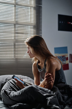 Offended female teenager in grey vest sitting on bed against window covered with venetian blinds while suffering from cyberbullyingの写真素材