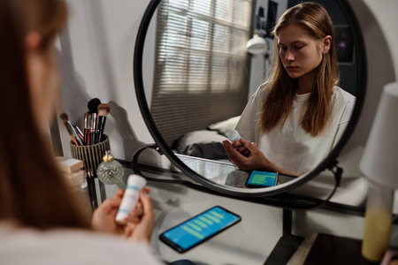 Reflection in mirror of stressed teenage girl looking at bottle of pills in her hands while...