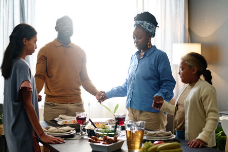 Young family of four holding by hands while standing around served table with homemade food and...