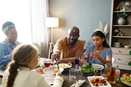 Young man putting corn on plate of his daughter over served festive table with homemade drinks and food while sitting among family membersの写真素材