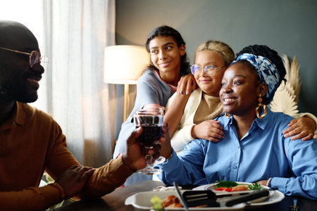 Happy young African American couple holding glass of red homemade wine and looking at one another while sitting by table with daughtersの写真素材