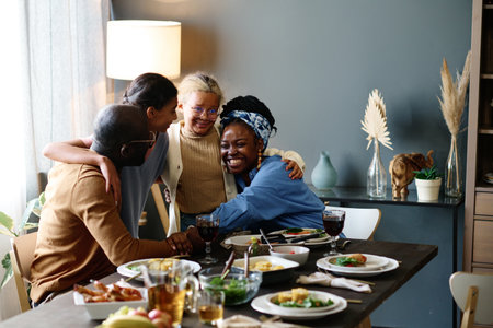 Cheerful young woman embracing one of two daughters by festive table served with homemade food and drinks during Thanksgiving dinnerの写真素材