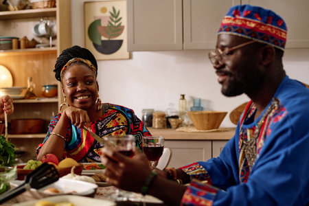 Happy African American woman in ethnic apparel looking at her husband with smile while sitting in front of him by served festive table at homeの写真素材