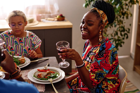 Happy young African American woman in ethnic attire holding wineglass with red homemade wine during celebration of national holidayの写真素材