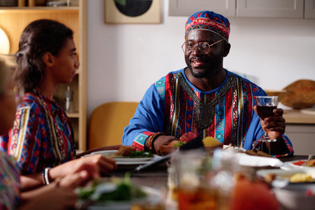 Focus on young African American man with glass of red homemade wine sitting by served festive table and talking to his daughters by dinnerの写真素材