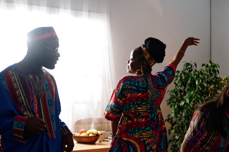 Young African American woman in ethnic attire keeping arm raised during dance while standing next to her husband wearing national costumeの写真素材