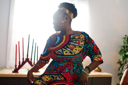 Young African American woman in ethnic attire and accessories performing national dance by table...