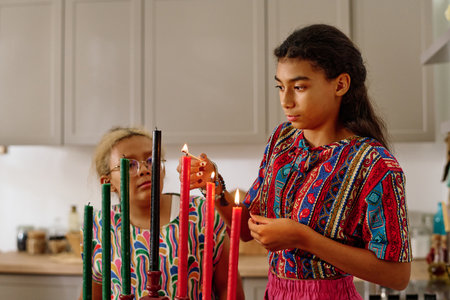 Cute African American girl in national attire burning candles for kwanzaa celebration while standing in front of camera against younger sisterの写真素材