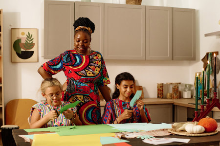 Young African American woman in national attire looking at her daughters cutting paper while preparing greeting cards for celebration of kwanzaaの写真素材