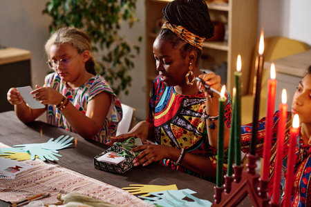Focus on young woman in national costume packing kwanzaa gifts for her family while sitting next to her daughter burning seven candlesの写真素材