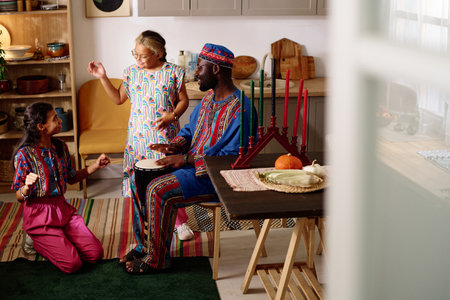Happy young African American man beating drum and looking at his daughters in national costumes performing dance at kwanzaa home partyの写真素材