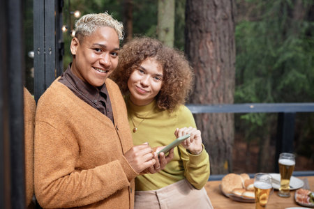 Portrait of girls smiling at camera while watching photos on smartphone during picnic outdoorsの写真素材