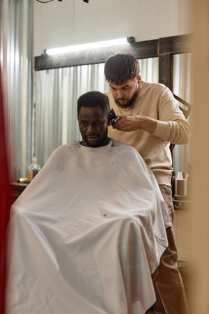 Vertical image of African American man sitting in chair while hairdresser cutting his hair with haircut machineの写真素材
