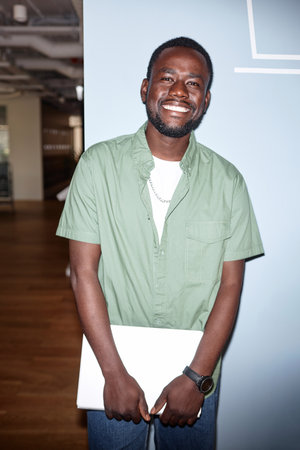 Vertical image of African American man with laptop smiling at camera while standing in office hallの写真素材