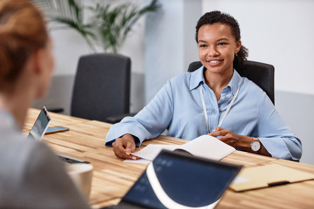 Young smiling businesswoman discussing work with her colleague while they sitting at table at meetingの写真素材