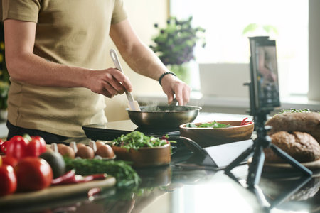 Medium shot of unrecognizable man standing by stove at kitchen frying food in pan while being filmed on smartphoneの写真素材