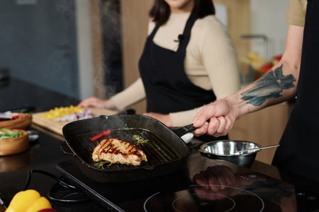Closeup of unrecognizable man holding pan with prepared piece of meat showing it to camera with unknown woman standing on background, focus on meatの写真素材