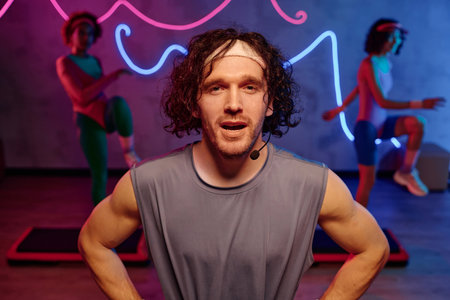 Caucasian fitness instructor speaking into headset mic with young women training on backgroundの写真素材