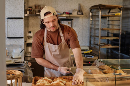 Bakery worker putting croissants from iron tray to wicker basket for sale