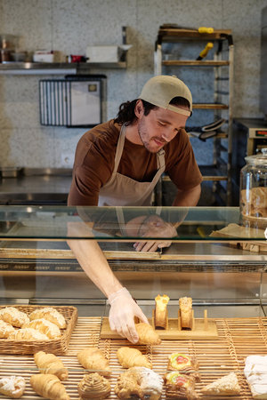 Bakery worker putting pastry assortment on wicker tray at showcase