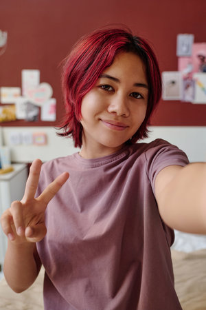 Cute smiling Asian female teenager with peace gesture looking at you while making video about preparation for plastic operation in clinicsの写真素材