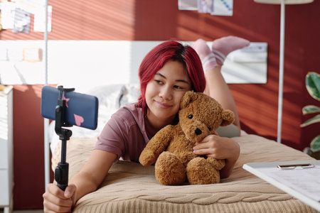 Cute teenage girl with teddybear relaxing on hospital bed in ward during online communication with subscribers of her channelの写真素材
