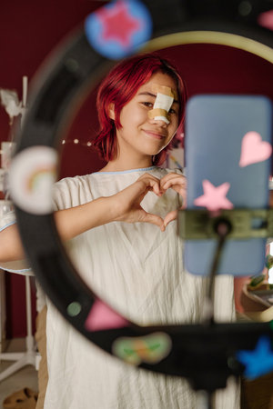 Happy teenage girl with bandage on her nose making heart shape with hands in front of smartphone...