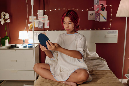 Teenage girl with liftmarks on her face sitting on bed in hospital ward and looking in mirror before cosmetic procedure or plastic surgeryの写真素材