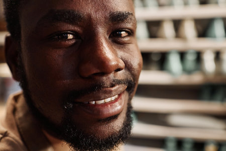 Face of happy young bearded African American craftsman, master or apprentice of shoemaker looking at camera in workshopの写真素材
