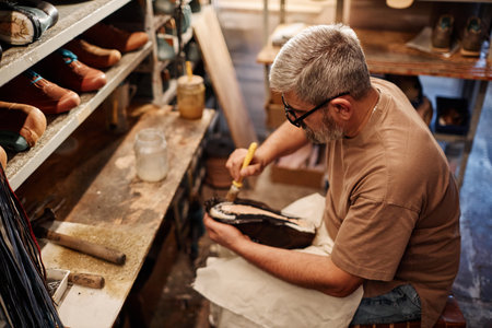 Side view of mature experienced shoemaker spreading glue over sole of unfinished boot or shoe while...