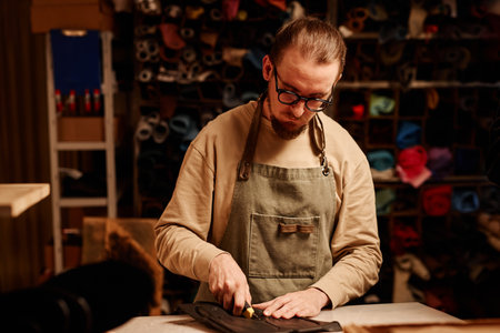 Young serious shoemaking master in eyeglasses and workwear cutting out sole workpieces by workplace...